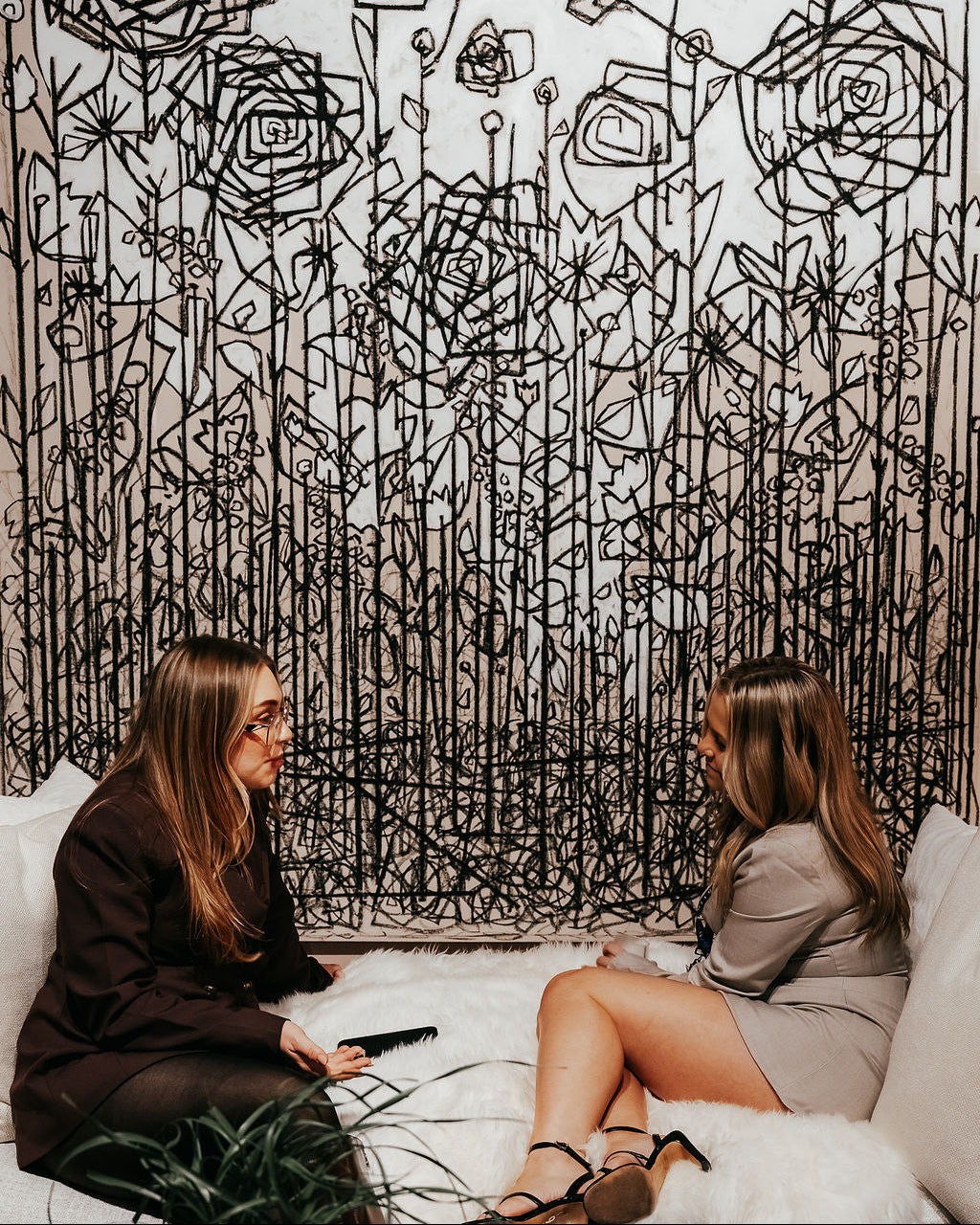 Two women sitting on a couch in front of a large abstract black and white floral artwork.